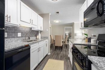 Galley kitchen with black appliances, white cabinets, wood-look flooring and grey tile backsplash.